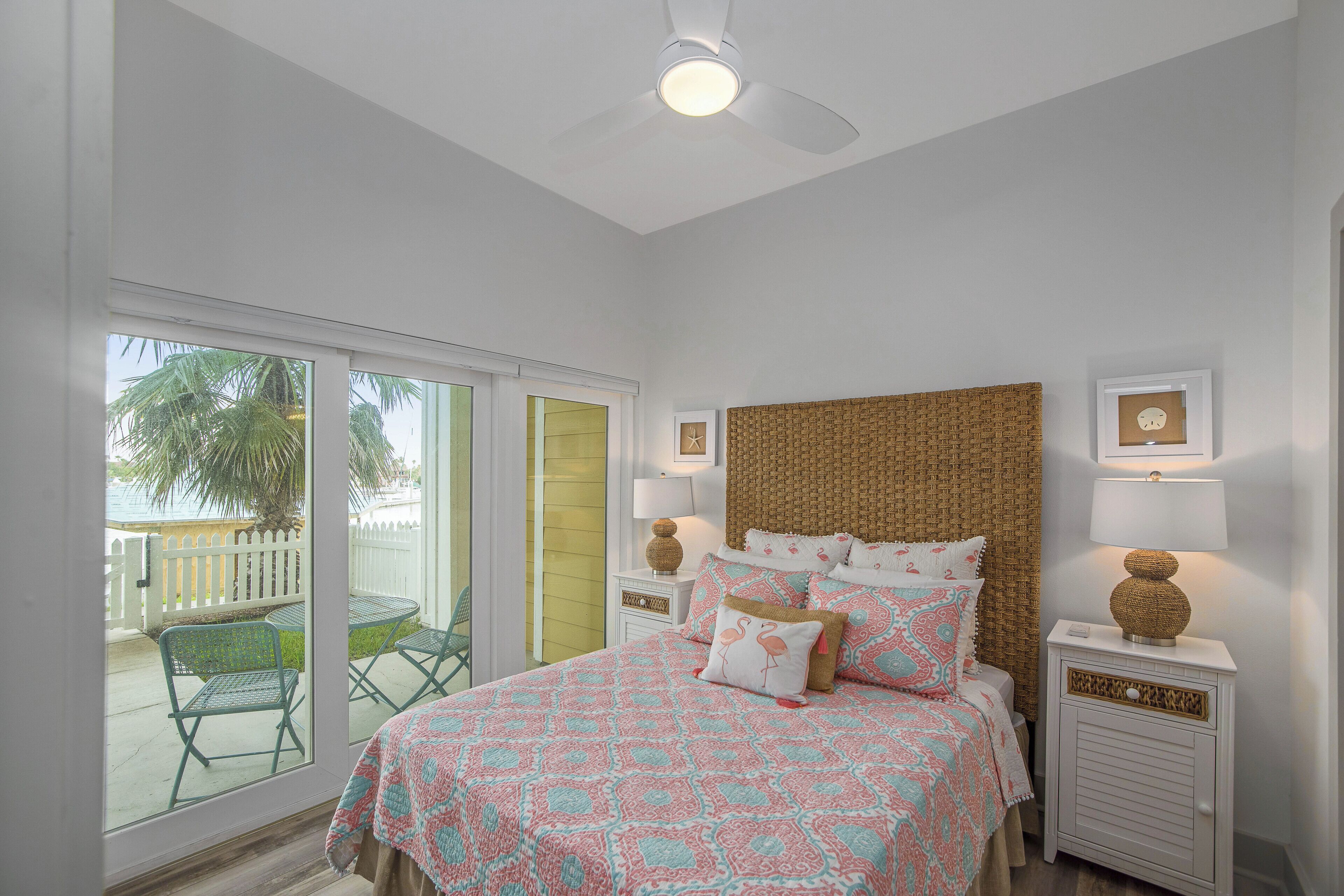 A bedroom with a bed, two nightstands, a ceiling fan, and a view of a balcony with chairs and a palm tree.