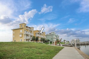 A multi-story residential building with a grassy area in front and a dock extending into a body of water. - Marina Del Bell - Elevator! Boat Slip for Rent! (Port Aransas)