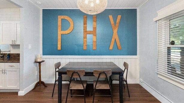 A dining area with a table and chairs, a wall-mounted sign with 'PHX' letters, a chandelier, and a window with blinds. - Centra Phoenix House Kingbed (Phoenix)