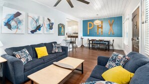A modern living room with a dark blue sofa, yellow cushions, a wooden coffee table, abstract wall art, a ceiling fan, and a dining area with a table and chairs. - Centra Phoenix House Kingbed (Phoenix)