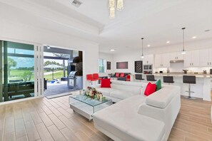 Una sala moderna con una mesa de centro de vidrio, un sofá blanco con cojines rojos y verdes, y una zona de cocina con armarios blancos y una barra.