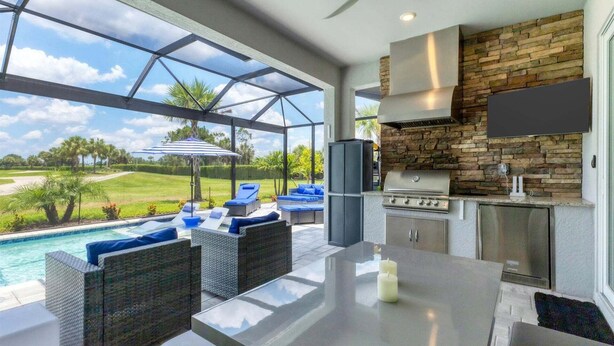 A modern outdoor kitchen with a stone backsplash, a large TV mounted above the counter, and a poolside seating area with wicker furniture. - Heritage Landing Haven Resort Amenities + Golf Membership (Punta Gorda)