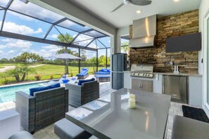 Eine moderne Außenküche mit Steinrückwand, einem großen Fernseher über der Arbeitsplatte und einem Sitzbereich am Pool mit Korbmöbeln.