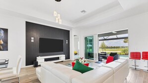 A modern living room with a large flat-screen TV, white sofas, a glass coffee table, and a view of a golf course through large sliding glass doors. - Heritage Landing Haven Resort Amenities + Golf Membership (Punta Gorda)