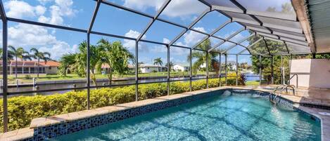 A clear swimming pool with a glass enclosure, surrounded by greenery and a view of a residential area.