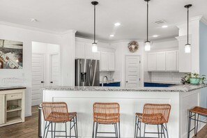 A kitchen with white cabinetry, a stainless steel refrigerator, a granite countertop, and three wicker bar stools. - Sunrise Paradise - Pet Friendly! Boat Parking! (Port Aransas)