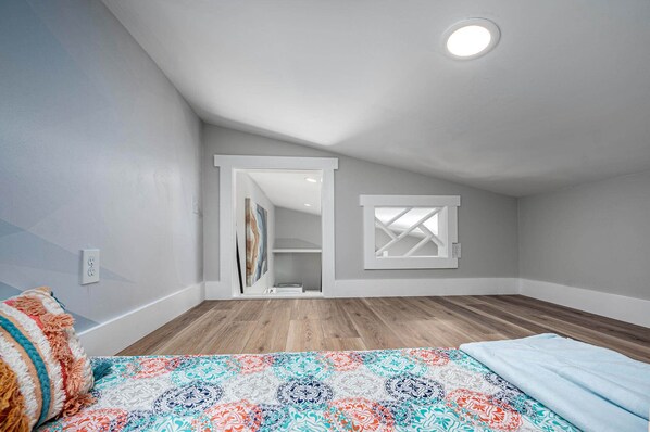 Une chambre avec un plafond en pente, un plancher en bois, un lit recouvert d’une courtepointe colorée, une petite fenêtre et une porte donnant sur une autre pièce.