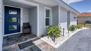 A house with a blue door, a welcome mat, a wicker chair, and a small garden with plants and a pathway.