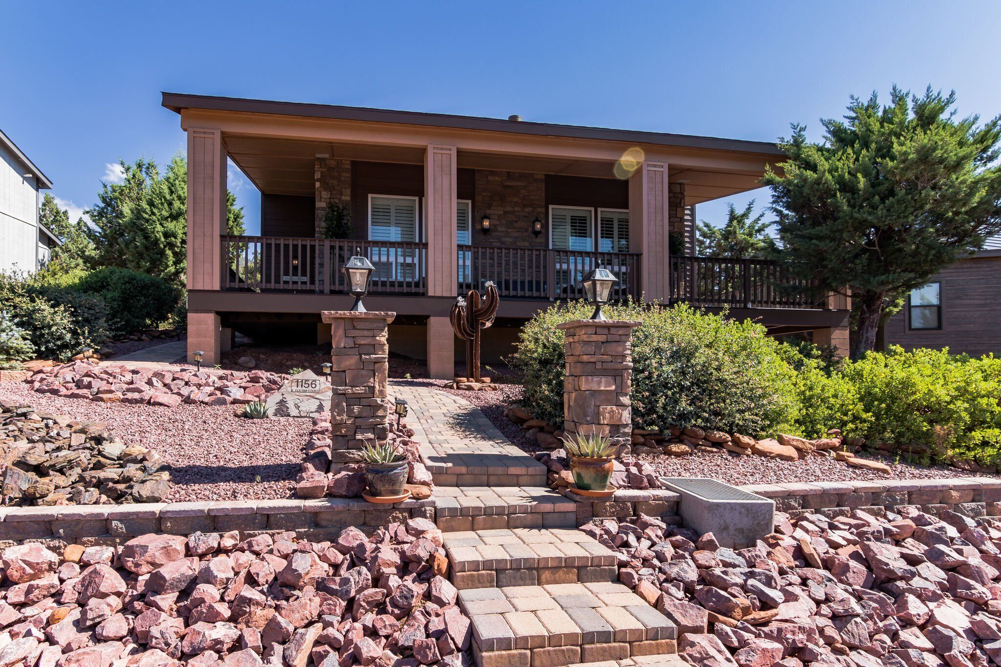 A two-story house with a covered porch, stone pillars, and a landscaped front yard with rocks and plants.