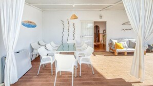 Een moderne kamer met een glazen tafel, omringd door witte stoelen, een houten vloer en een bank met een geel kussen.