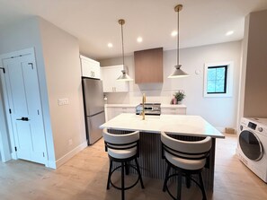A modern kitchen with a central island, bar stools, pendant lights, a built-in oven, and a washing machine. - Modern 1BR Suite, 5 Min to UVIC and Gyro Beach (Victoria)