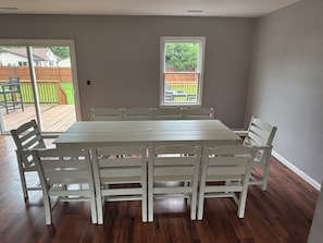 A dining area with a rectangular table and six chairs, large windows with a view of a backyard, and a wooden deck.