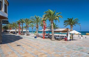 Un patio frente a la playa con palmeras, mobiliario exterior y un restaurante bar.