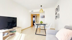 Uma sala de estar moderna com uma televisão de tela plana, um sofá cinza, uma mesa de jantar com cadeiras e uma cozinha visível através de uma porta aberta.