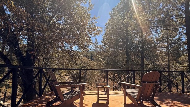 Un patio al aire libre con sillas y mesa de madera, rodeado de árboles y una barandilla de metal.