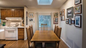 Une cuisine avec des meubles en bois, une cuisinière et un coin repas comprenant une table et des chaises. Il y a une lucarne au-dessus de la cuisine et une fenêtre avec des stores.