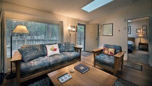 Un salon chaleureux avec un canapé et un fauteuil, une table basse et une lampe. Il y a des fenêtres de toit au plafond et une porte qui donne sur une autre pièce.