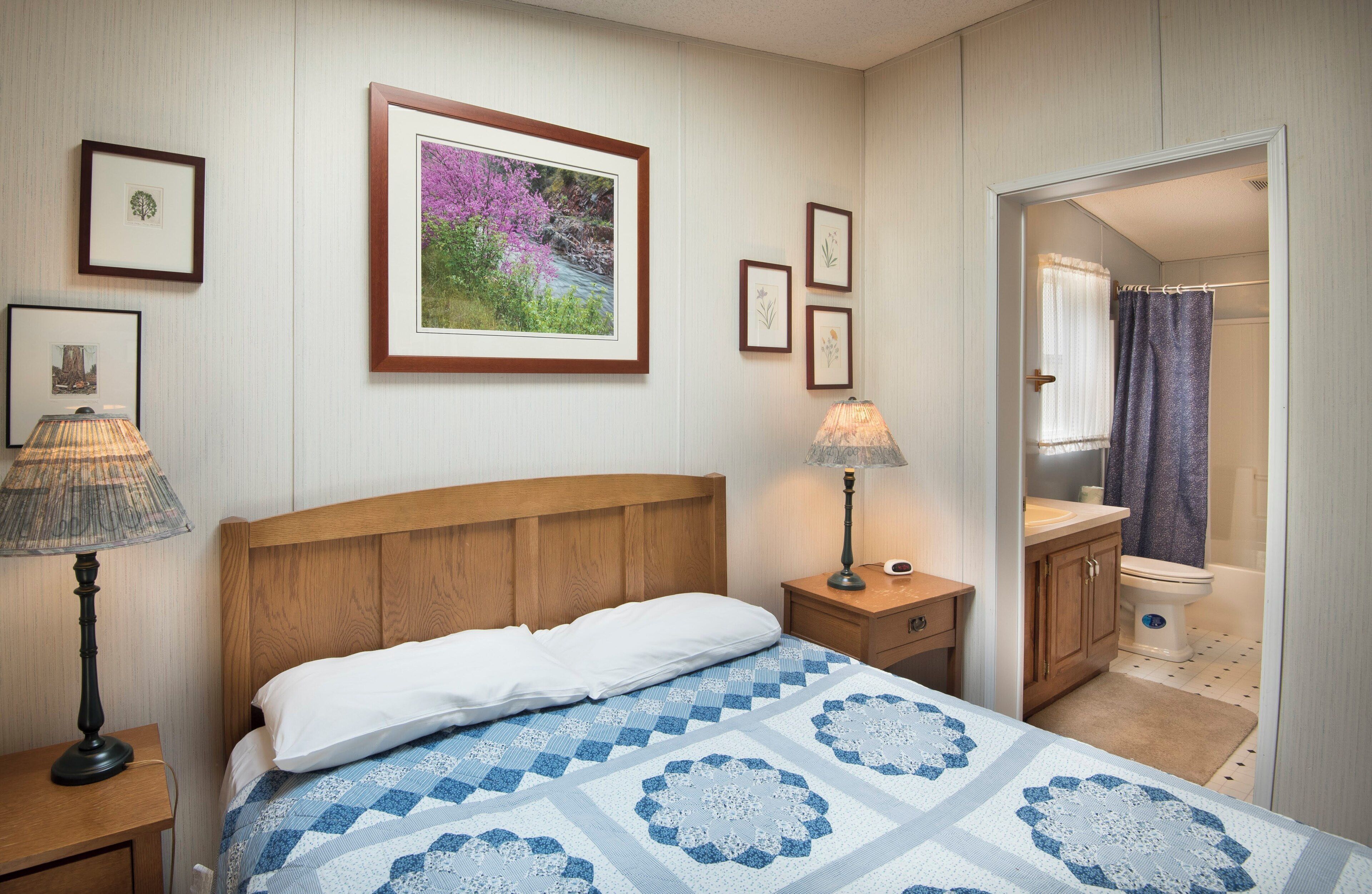 A bedroom with a wooden headboard, a bed with a blue and white patterned bedspread, two bedside lamps, a nightstand, a framed picture on the wall, and a bathroom visible through an open door.