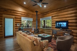 Interno di una baita di legno con divano, poggiapiedi, televisore e camino.