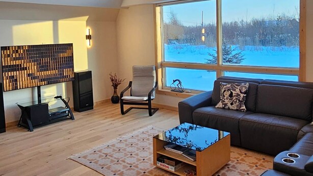 Un salon spacieux avec un plafond haut, de grandes fenêtres, un plancher en bois, un canapé en cuir noir, une table basse en verre et un haut-parleur fixé au mur.