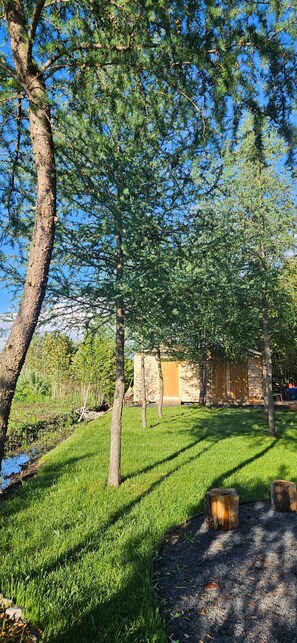 A stone building with a grassy yard, trees, and a small stream. - Modern Forest Retreat with Sauna, Hot Tub, Fireplace (Rural Municipality of Reynolds)