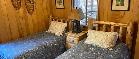 Una habitación tipo cabaña de madera con dos camas, una mesita de noche con lámpara y cuadros en las paredes.