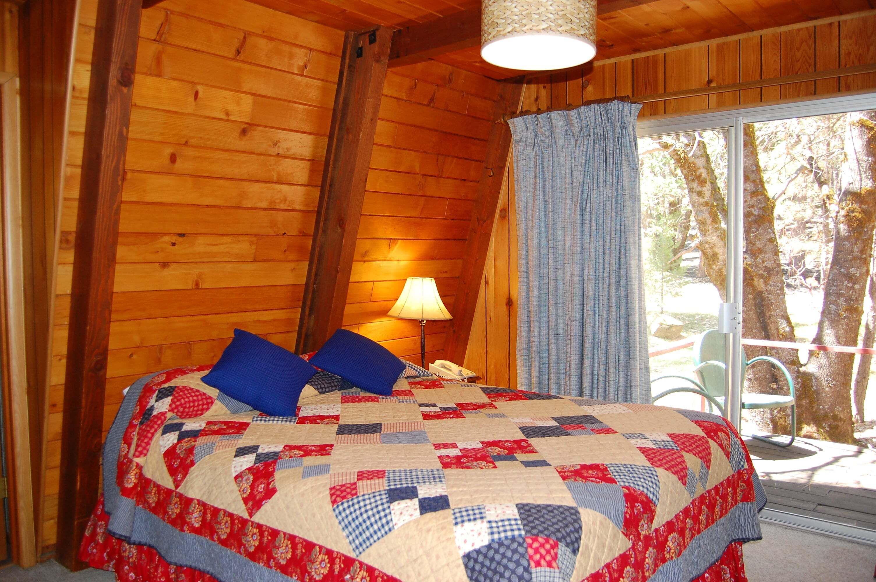 Camera da letto in una baita di legno, con copripiumino trapuntato, una lampada e vista sugli alberi attraverso una finestra.