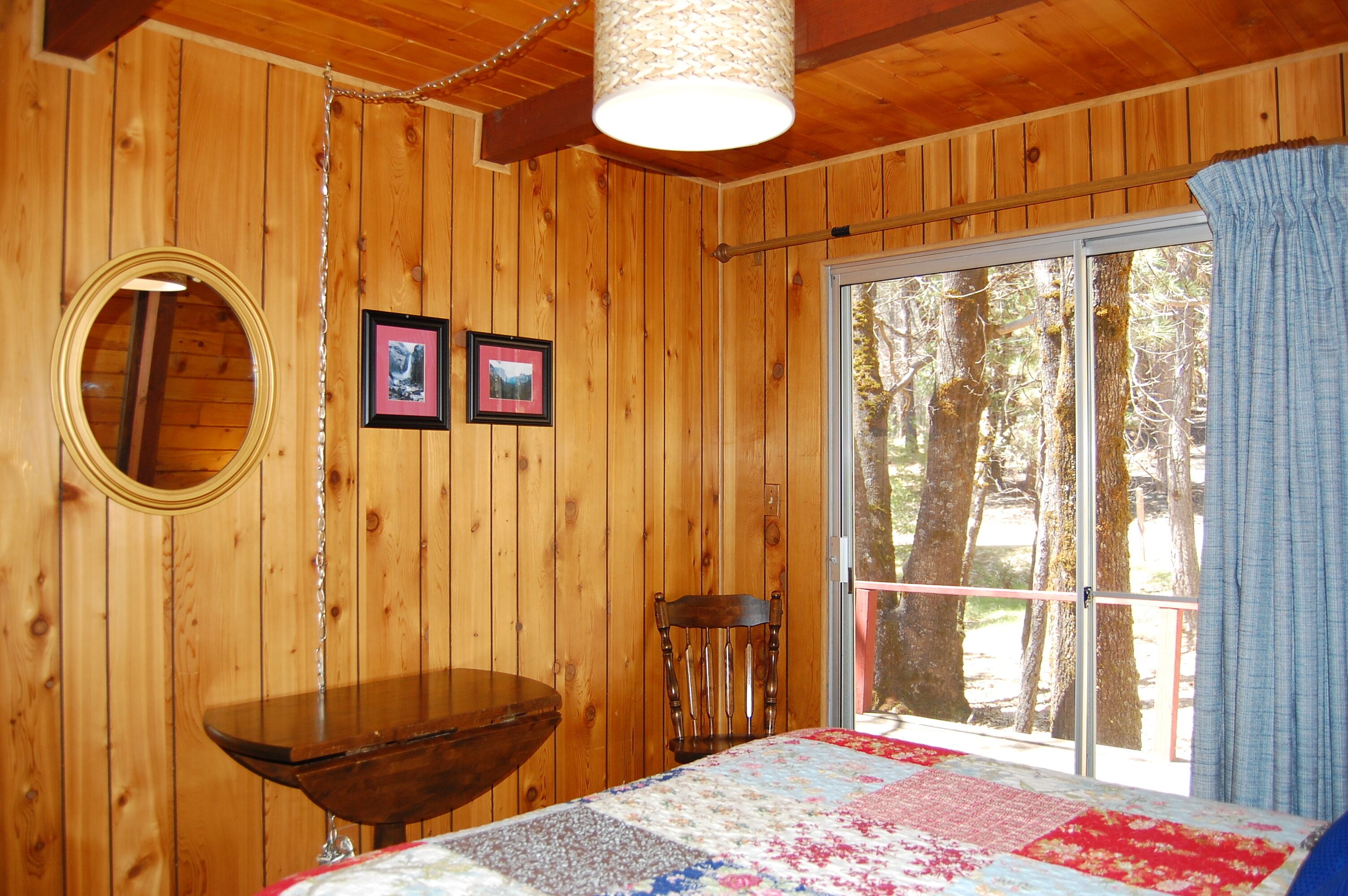 Ein Zimmer in einer Holzhütte mit einem Bett, einem Tisch, einem Stuhl, einem Fenster mit Blick auf Bäume und zwei gerahmten Bildern an der Wand.