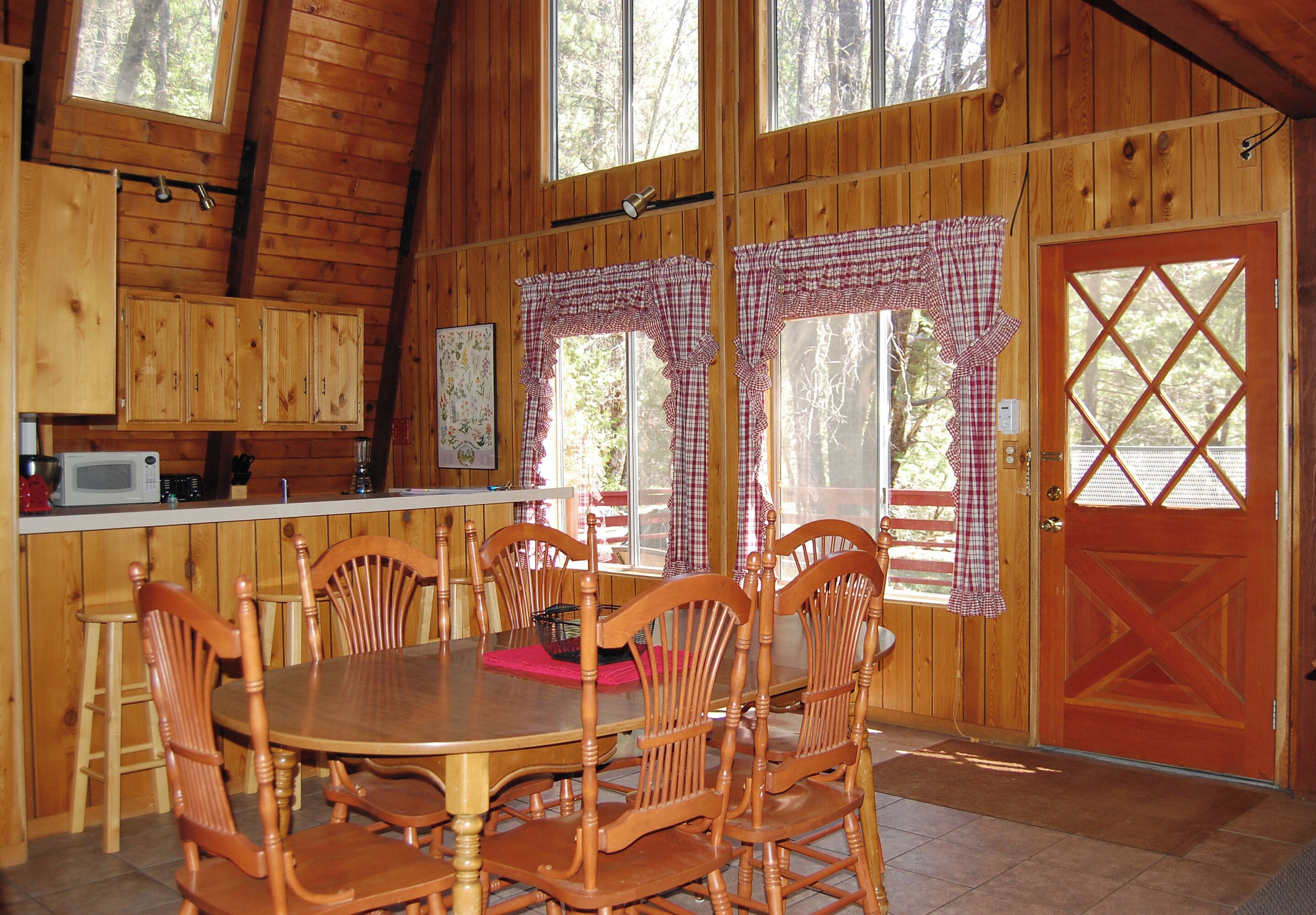 A wooden cabin interior with a dining table, chairs, a kitchen area, and large windows with checkered curtains.