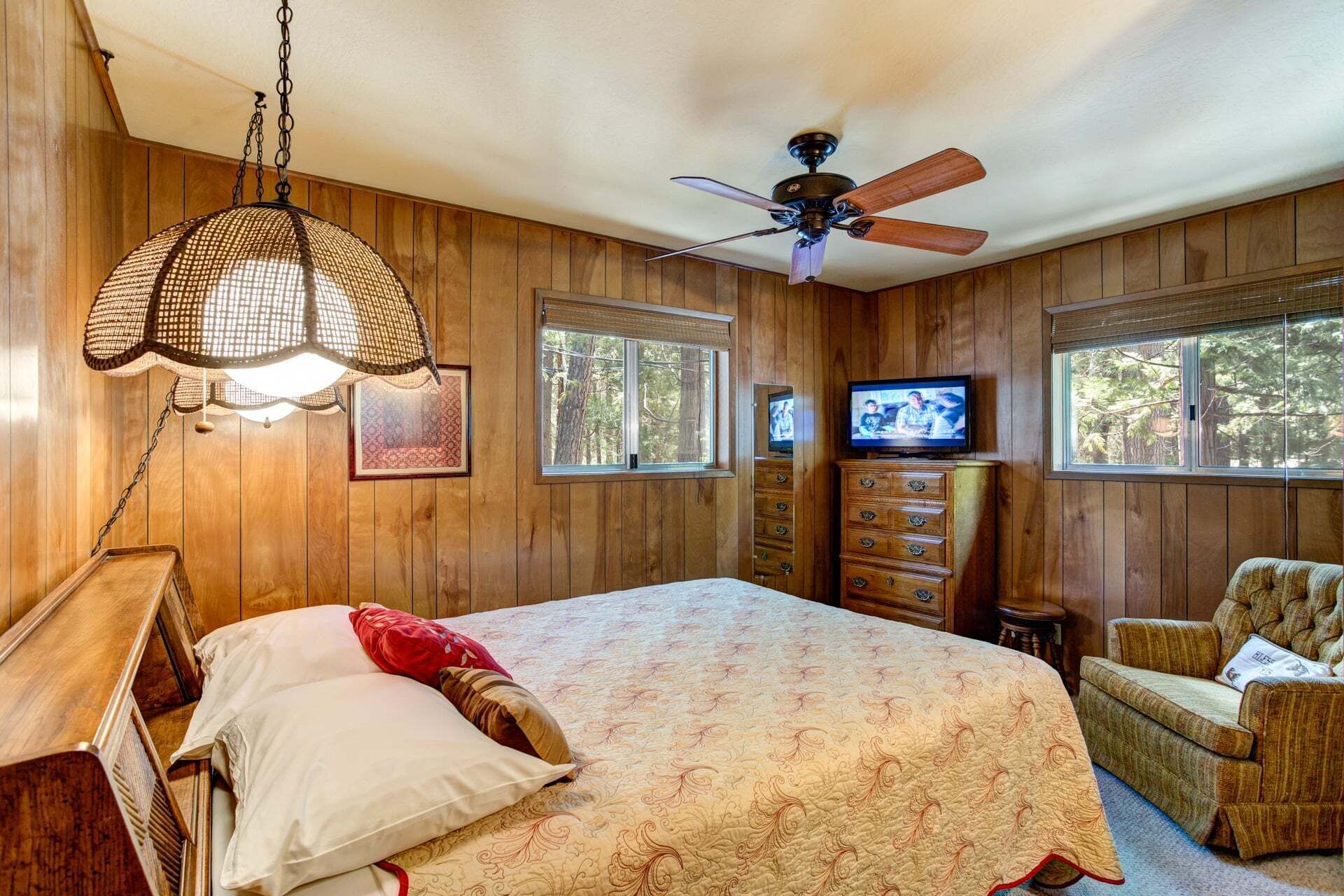 A wooden-paneled bedroom with a bed, a ceiling fan, a television, and a window.