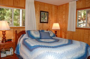 A wooden-paneled bedroom with a large bed, two bedside lamps, a nightstand with books, and a framed picture on the wall. - River Song (Yosemite National Park)