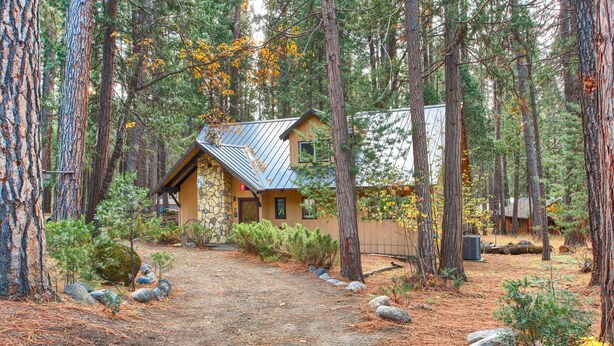 A rustic cabin with a stone foundation and a metal roof, surrounded by dense forest and a gravel path. - Spindler's Chalet (Yosemite National Park)
