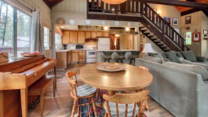 A spacious living area with a wooden piano, a round dining table, and a comfortable sofa. A staircase leads to an upper level with a balcony. - Spindler's Chalet (Yosemite National Park)