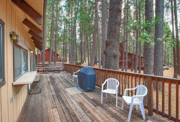 A wooden deck with two white chairs and a barbecue grill, overlooking a forested area with tall trees and a red cabin in the background. - Spindler's Chalet (Yosemite National Park)
