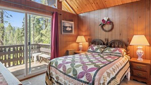 Un dormitorio con una cama grande, paredes de madera, techo de madera, vista a árboles, un banco y una mesa afuera de la ventana.