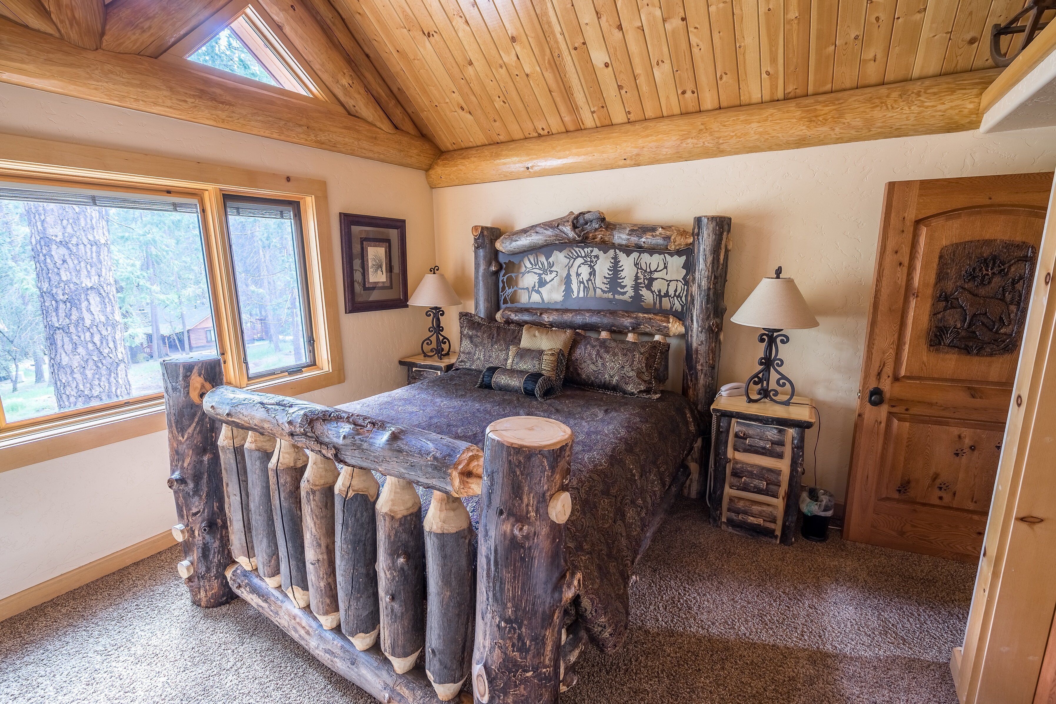 Une chambre à coucher au style rustique, dotée d’un grand lit avec une tête de lit en bois ornée de sculptures d’animaux, de deux lampes de chevet et d’une porte en bois avec un motif sculpté.