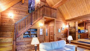 Interior de una cabaña de madera con una escalera, un sofá y una estantería.