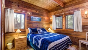 Un dormitorio de cabaña de madera con una cama, dos mesitas de noche, una silla y dos ventanas con cortinas.