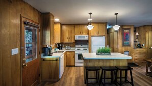 A kitchen with wooden cabinets, a white stove, a microwave, a refrigerator, and a coffee maker. There are three bar stools and a potted plant on the island. - Pine Creek Cabin (Yosemite National Park)