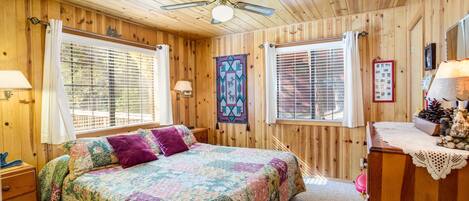A cozy wooden cabin bedroom with a bed, a dresser, a nightstand, a ceiling fan, and two windows with blinds.