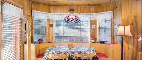 A wooden-paneled room with a round table, two chairs, a chandelier, and lace curtains.