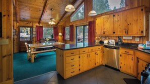 Interior de una cabaña de madera con cocina, comedor y un amplio ventanal con vistas a un bosque.