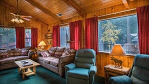 Interior acogedor de una cabaña con paredes y techos de madera, un ventanal con cortinas rojas y vistas a árboles.