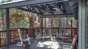 Eine überdachte Terrasse im Freien mit Holzmöbeln, darunter Schaukelstühle und Bänke, umgeben von Bäumen.