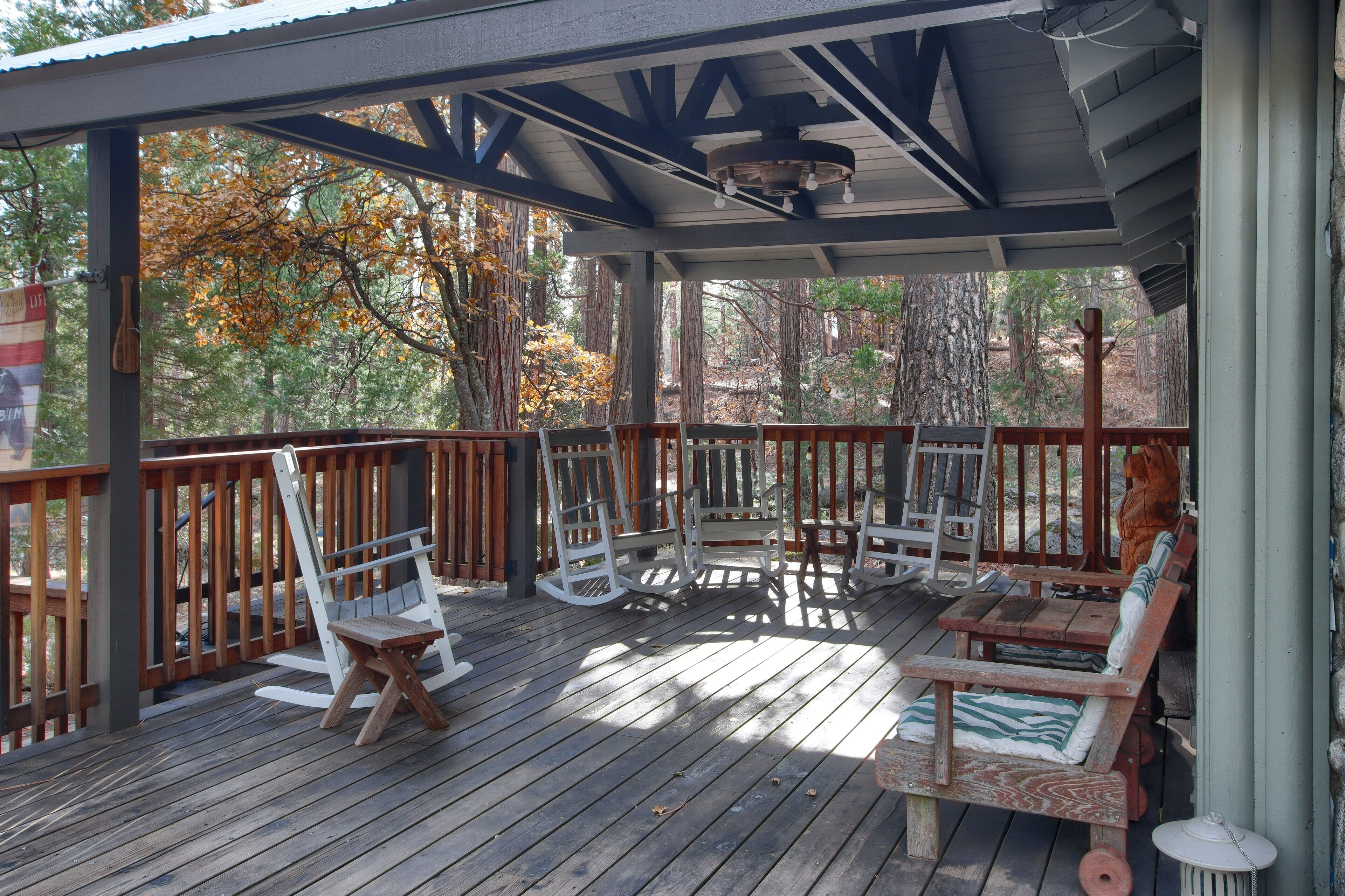 Une terrasse extérieure couverte, aménagée avec du mobilier en bois, notamment des fauteuils à bascule et des bancs, et entourée d’arbres.