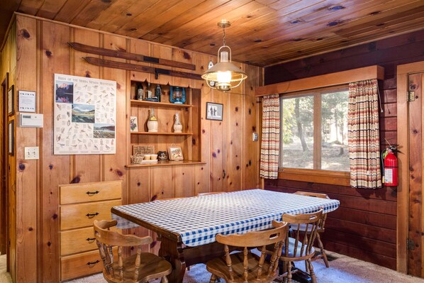 L’intérieur d’un chalet en bois comprend une table à manger, des chaises, un luminaire suspendu, une fenêtre avec des rideaux et un extincteur fixé au mur.