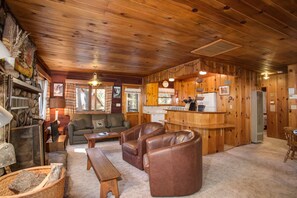 L’intérieur chaleureux d’un chalet, avec des murs et un plafond en bois, un salon doté d’un canapé et de fauteuils en cuir, un coin cuisine avec un réfrigérateur et une cuisinière, et une cheminée avec des bûches.