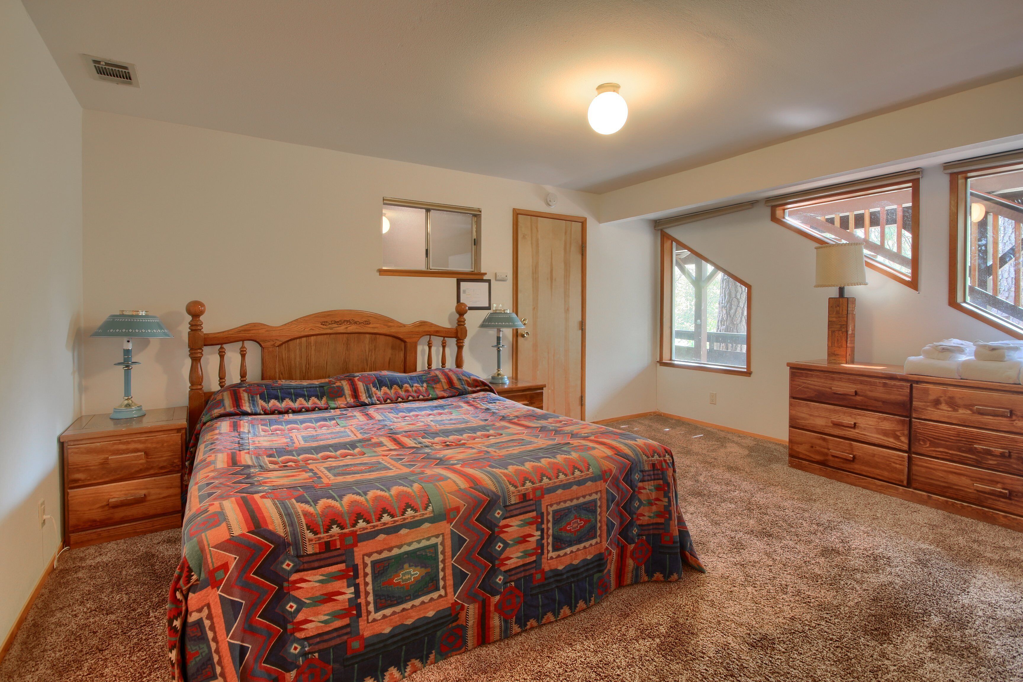 A bedroom with a large bed, wooden headboard, two bedside tables with lamps, a dresser, and a window with a view of trees.