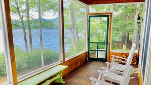 A covered deck with wooden flooring, white rocking chairs, a green bench, and large windows overlooking a body of water. - 30fl Amazing Lake House 20 min From Bretton Woods Stunning Water Views! Bonus Room (Whitefield)