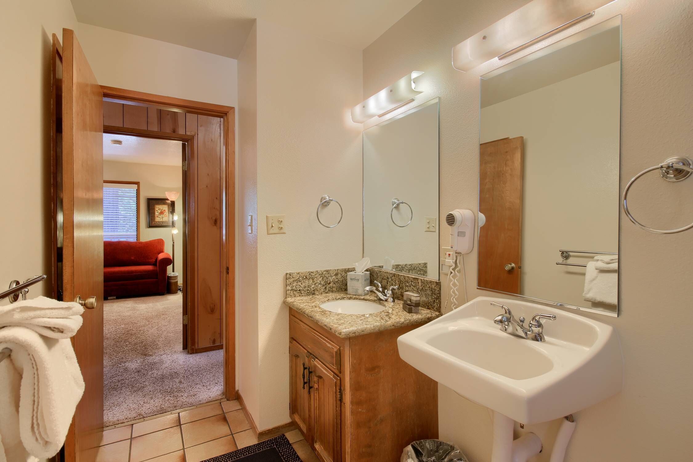 A bathroom with a sink, mirror, and towel rack. There is a door leading to a living area with a red couch.